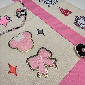 May include: A cream-colored canvas tote bag with pink fabric straps, decorated with various embroidered patches including a pink bow, a Minnie Mouse head, a castle, and a cat. The bag features a beaded strap and a black flower-shaped brooch.