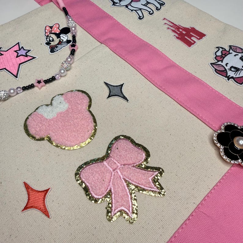 May include: A cream-colored canvas tote bag with pink fabric straps, decorated with various embroidered patches including a pink bow, a Minnie Mouse head, a castle, and a cat. The bag features a beaded strap and a black flower-shaped brooch.