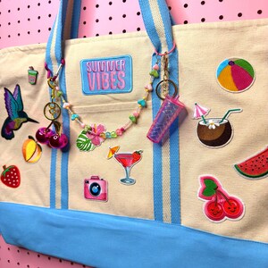 May include: A cream-colored tote bag with blue stripes and handles, adorned with colorful patches of summer-themed items. The bag features the text "SUMMER VIBES" and is accessorized with charms and a beaded strand.
