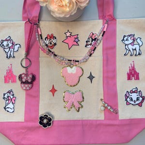 May include: A beige and pink canvas tote bag decorated with various patches including Marie the cat, pink castles, Minnie Mouse, stars, and bows. The bag features a beaded necklace, a Minnie Mouse keychain, and a black flower pin attached to the front.