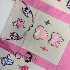 May include: A cream-colored canvas tote bag with pink fabric trim, decorated with embroidered patches of Minnie Mouse, a pink bow, a castle, and a cat. A beaded lanyard with black and pink beads and a Minnie Mouse keychain are attached.