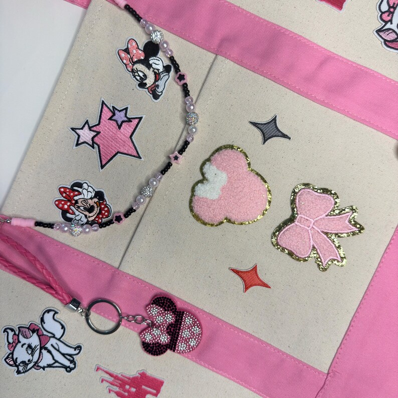May include: A cream-colored canvas tote bag with pink fabric trim, decorated with embroidered patches of Minnie Mouse, a pink bow, a castle, and a cat. A beaded lanyard with black and pink beads and a Minnie Mouse keychain are attached.