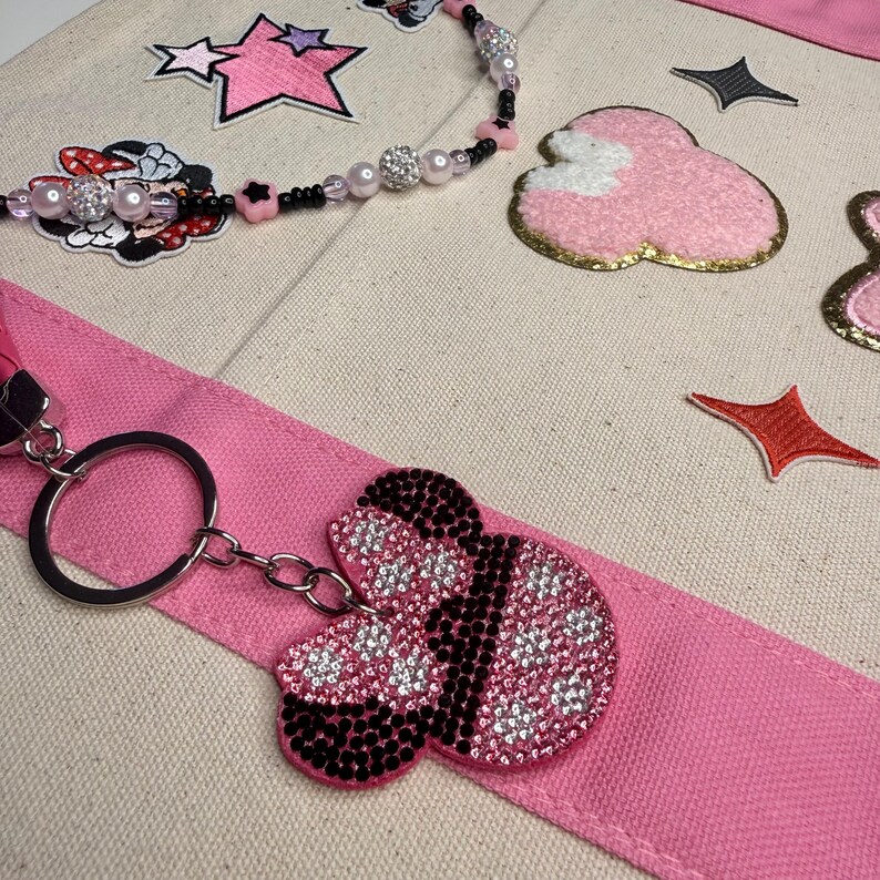May include: A beige canvas tote bag featuring a pink fabric strap, a beaded lanyard with black and pink star beads, and various embroidered patches including stars, a pink cloud, and a rhinestone-covered keychain shaped like a mouse head.