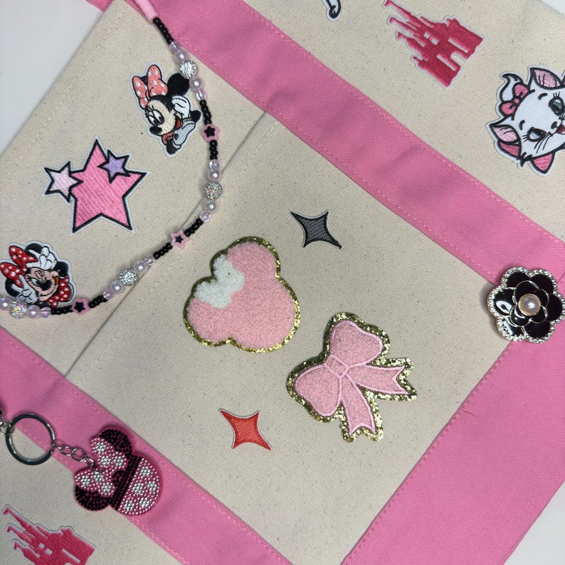 May include: A cream-colored canvas tote bag with pink fabric trim, decorated with various embroidered patches including a pink bow, a textured pink shape, cartoon characters, and a castle. A beaded lanyard and a keychain are attached.