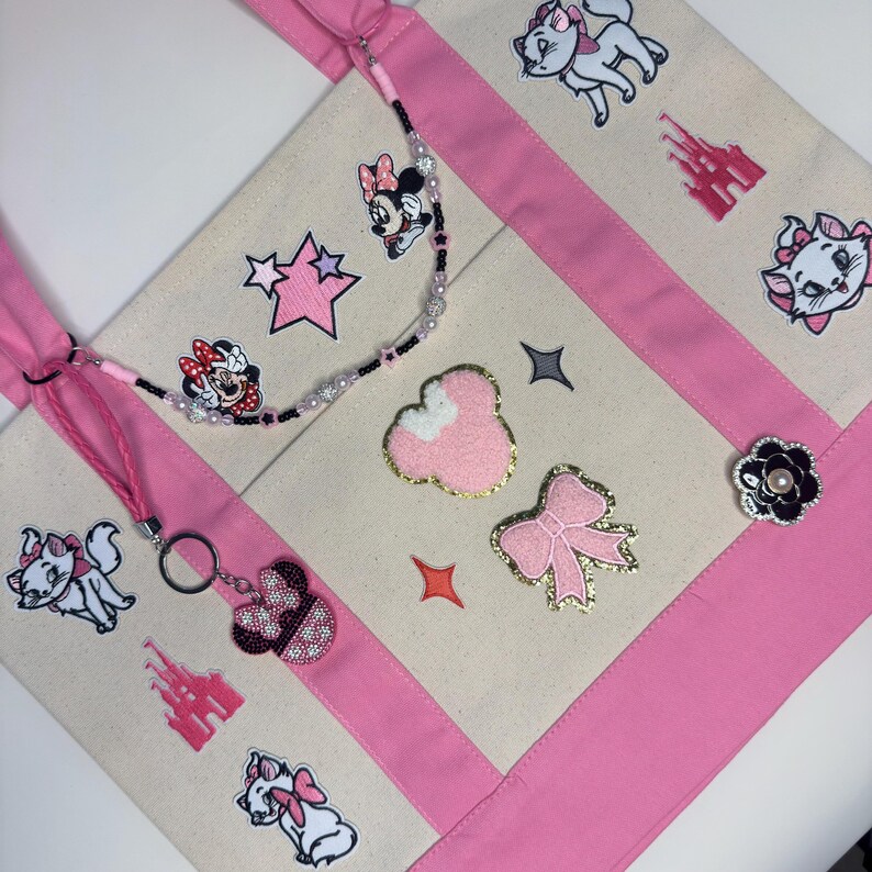 May include: A cream-colored canvas tote bag with pink fabric straps and trim. The bag features embroidered patches of white kittens, pink castles, and Minnie Mouse, along with a beaded strap accessory and a pink glittery bow and ear patch.