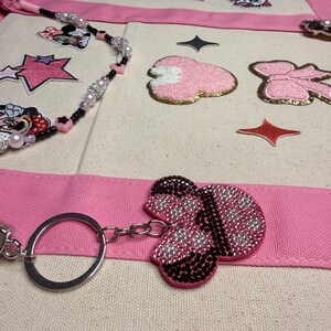 May include: A cream-colored canvas tote bag featuring pink fabric straps, decorated with embroidered patches of a cartoon character, pink bows, and stars. A beaded strap with black, pink, and clear beads and a rhinestone-covered bow keychain are attached.