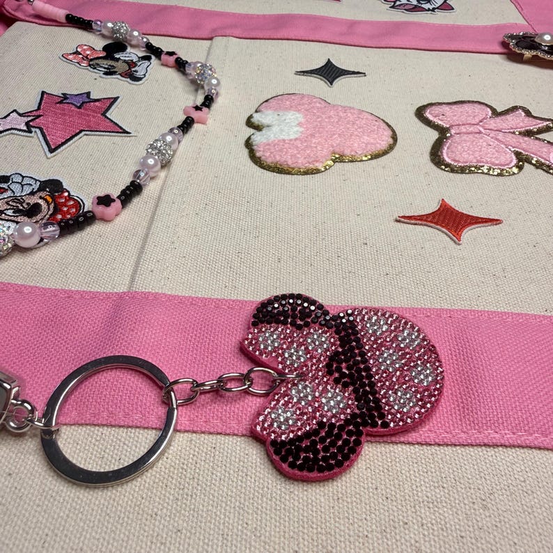 May include: A cream-colored canvas tote bag featuring pink fabric straps, decorated with embroidered patches of a cartoon character, pink bows, and stars. A beaded strap with black, pink, and clear beads and a rhinestone-covered bow keychain are attached.