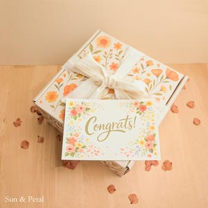 May include: A rectangular gift box decorated with an orange and yellow floral pattern, tied with a white fabric ribbon. A white rectangular card rests on top, featuring a colorful floral border and the gold script text Congrats!.