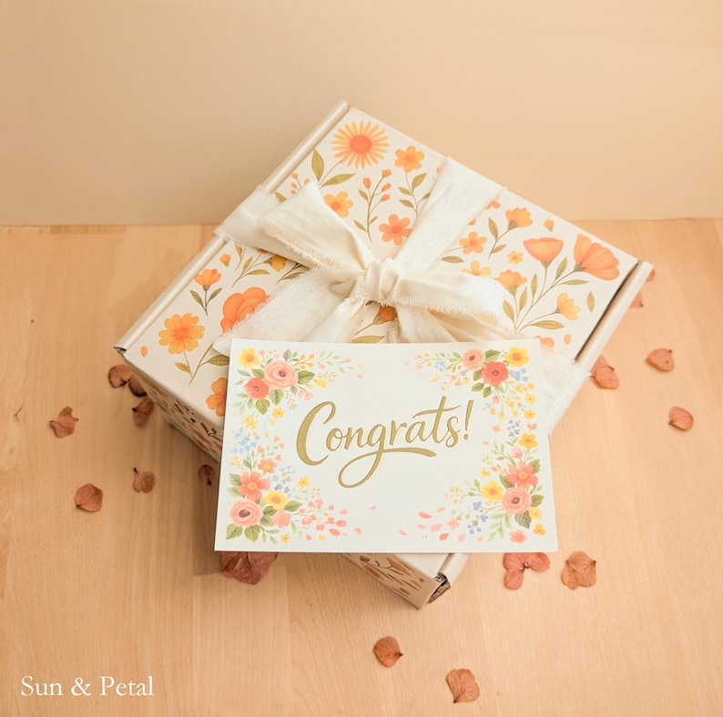 May include: A rectangular gift box decorated with an orange and yellow floral pattern, tied with a white fabric ribbon. A white rectangular card rests on top, featuring a colorful floral border and the gold script text Congrats!.