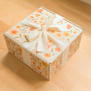 May include: A square cardboard box featuring a repeating pattern of orange and yellow flowers with green stems and leaves. The box is tied with a wide, off-white fabric ribbon in a bow on top. The text Sun & Petal is printed in the bottom corner.