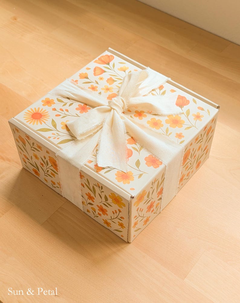 May include: A square cardboard box featuring a repeating pattern of orange and yellow flowers with green stems and leaves. The box is tied with a wide, off-white fabric ribbon in a bow on top. The text Sun & Petal is printed in the bottom corner.