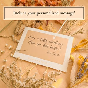 May include: A rectangular white greeting card with a tan center panel featuring the black cursive text: Here's a little something Hope you feel better! -Love Sarah. The card includes the text 2026 SUN & PETAL Co. and is surrounded by dried flowers.