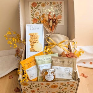 May include: A gift box decorated with floral patterns containing a Rustic Bakery cookie box, a ribbed ceramic mug with a yellow ribbon, three Tea With Tae tea sachets, a small jar of honey, and a dried flower bouquet with a thank you card.
