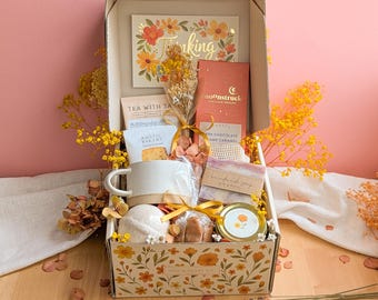 May include: A gift box decorated with orange and yellow flowers containing a Moonstruck dark chocolate bar, a Rustic Bakery cookie, a ceramic mug, a bar of soap, a small jar, a bath bomb, and a card with the text Thinking of You.