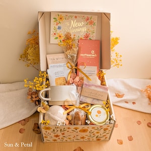 May include: A gift box featuring a card that reads To New Beginnings, a Moonstruck dark chocolate bar, a Rustic Bakery salted caramel cookie, a ceramic mug, a bar of handmade soap, a bath bomb, a small jar, and dried yellow flowers.