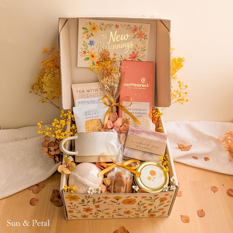 May include: A gift box featuring a card that reads To New Beginnings, a Moonstruck dark chocolate bar, a Rustic Bakery salted caramel cookie, a ceramic mug, a bar of handmade soap, a bath bomb, a small jar, and dried yellow flowers.