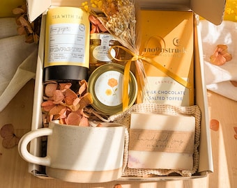 May include: A gift box containing a yellow Tea With Tae lemon ginger tea canister, a Moonstruck milk chocolate salted toffee bar, a Sun & Petal honey jar, a handmade soap bar in a mesh bag, a ceramic mug, and a bouquet of dried yellow flowers.