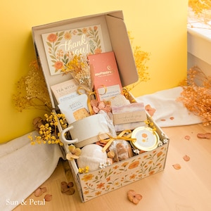 May include: A gift box featuring a thank you card, a Moonstruck dark chocolate bar, a speckled ceramic mug, a bar of handmade soap, a small gold-lidded jar, a bath bomb, and dried yellow flowers. The box exterior has a floral print and Sun & Petal text.