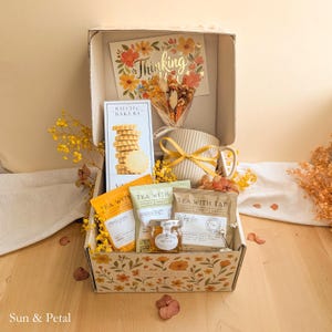 May include: A gift box containing a Rustic Bakery shortbread cookie package, a ribbed ceramic mug with a yellow ribbon, a small jar of honey, and two Tea With Tae tea bags. A floral card reading Thinking of You and dried flowers are included.