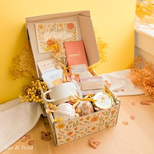 May include: A gift box decorated with orange floral patterns containing a ceramic mug, a Moonstruck dark chocolate bar, a small jar, a bar of soap, a bath bomb, and a Get Well Soon card. The box is filled with dried yellow flowers and tied with a ribbon.