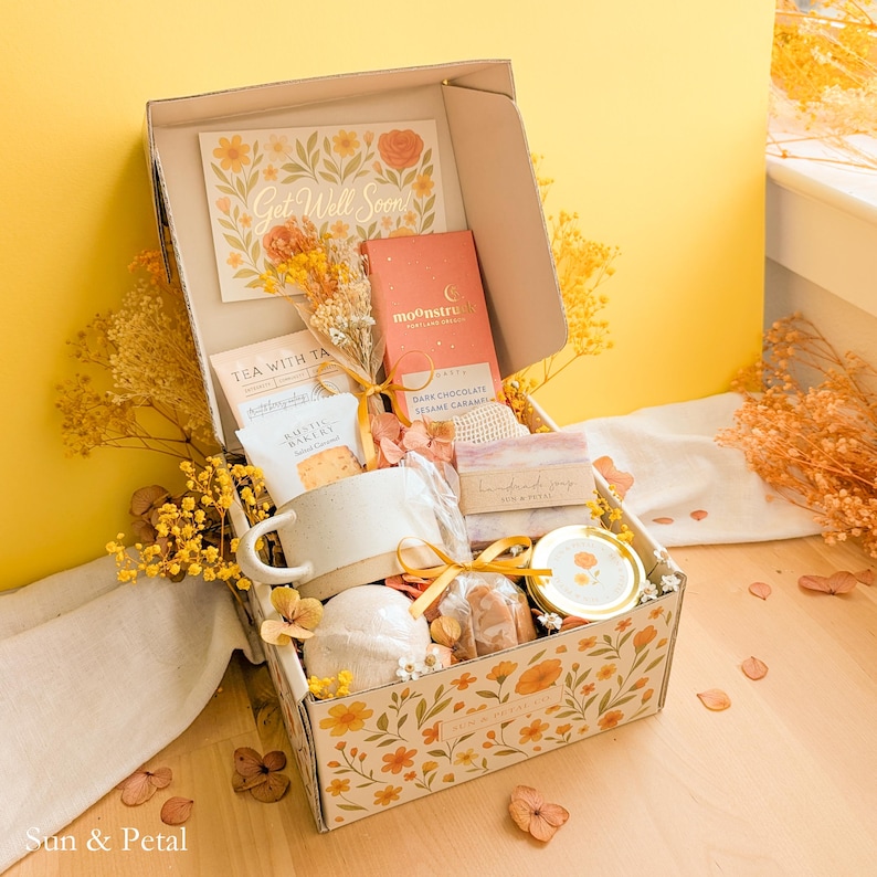 May include: A gift box decorated with orange floral patterns containing a ceramic mug, a Moonstruck dark chocolate bar, a small jar, a bar of soap, a bath bomb, and a Get Well Soon card. The box is filled with dried yellow flowers and tied with a ribbon.
