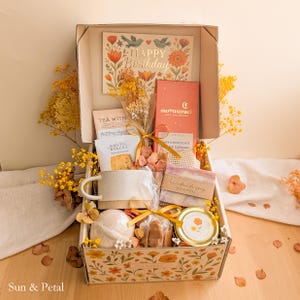 May include: A gift box filled with a variety of items, including a mug, soap, bath bomb, and a card that says "Happy Birthday." The box is decorated with floral patterns and contains a bouquet of dried flowers. The overall color scheme is warm, with shades of yellow and orange.
