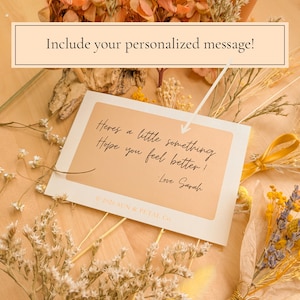 May include: A rectangular white greeting card with a tan center panel featuring the black cursive text: Here's a little something Hope you feel better! -Love Sarah. The card includes the text 2026 SUN & PETAL Co. and is surrounded by dried flowers.