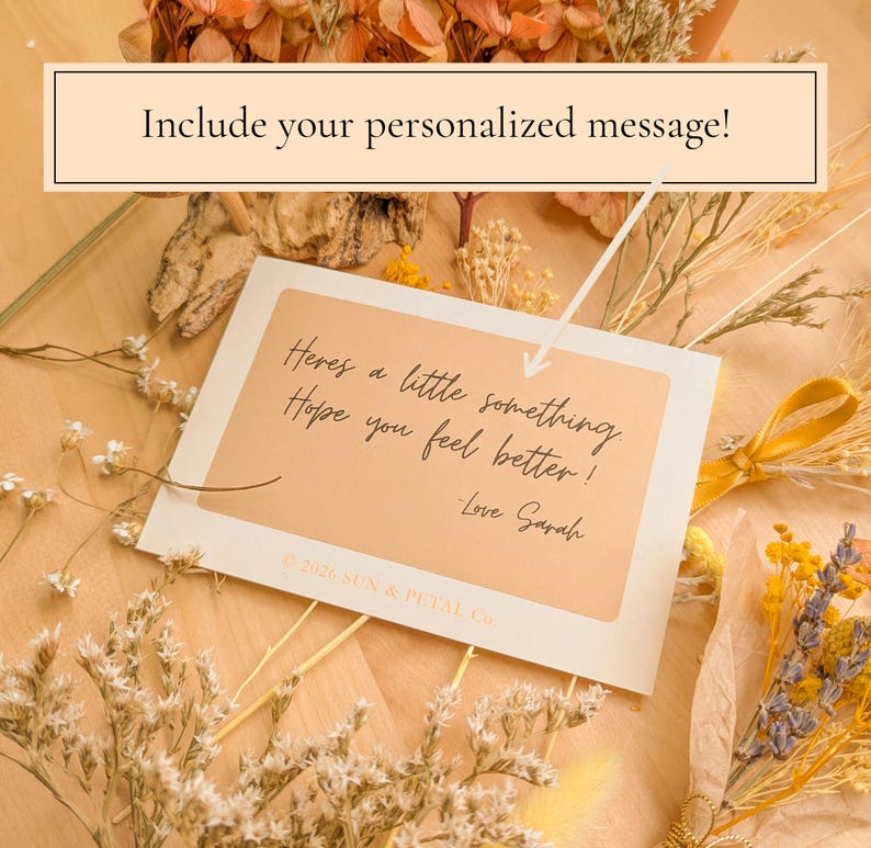May include: A rectangular white greeting card with a tan center panel featuring the black cursive text: Here's a little something Hope you feel better! -Love Sarah. The card includes the text 2026 SUN & PETAL Co. and is surrounded by dried flowers.