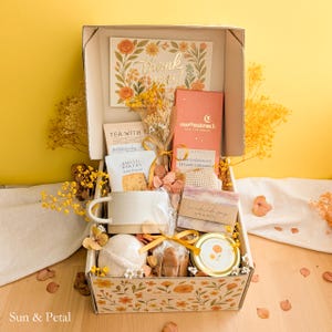 May include: A gift box containing a thank you card, a Moonstruck dark chocolate bar, a Rustic Bakery cookie, a ceramic mug, a bar of handmade soap, a bath bomb, and a small jar with a floral label, all decorated with dried yellow flowers.