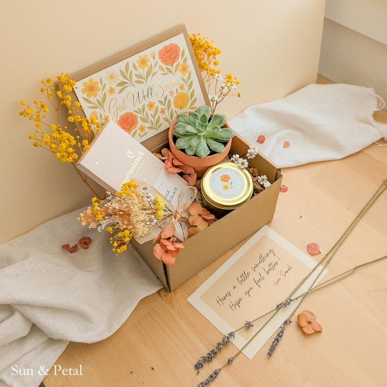 May include: A cardboard gift box containing a green succulent in a terracotta pot, a Moonstruck milk chocolate bar, a small candle with a gold lid, and dried yellow flowers. The box lid features a floral Get Well Soon card and a note.