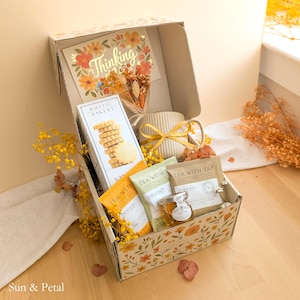 May include: A gift box decorated with orange and yellow flowers containing a Rustic Bakery cookie box, a ribbed ceramic mug with a yellow ribbon, three Tea With Tae tea packets, a small jar of honey, and a Thinking of You floral card.