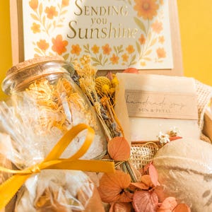 May include: A gift box containing a glass jar filled with yellow flower petals and bath salts, a bar of handmade soap labeled Sun & Petal, a round bath bomb, and a bundle of dried flowers. A card with the text Sending you Sunshine is included.