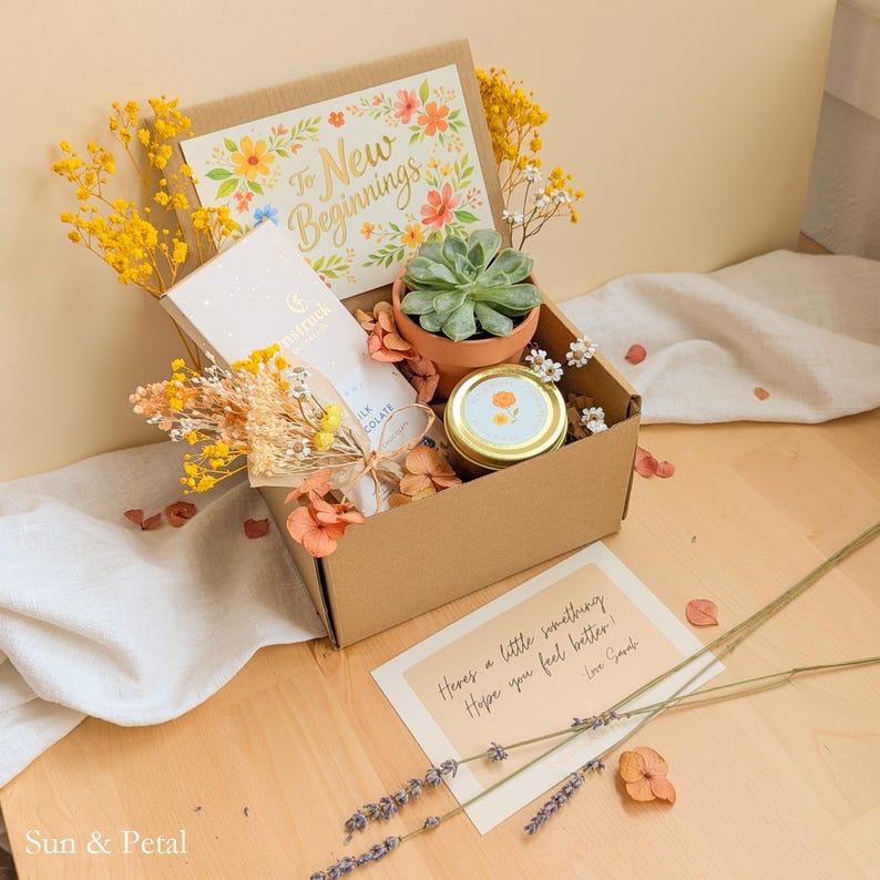 May include: A cardboard gift box containing a succulent in a terracotta pot, a gold-lidded candle, a chocolate bar, and dried yellow flowers. A card inside reads To New Beginnings, and a separate note reads Here's a little something Hope you feel better.