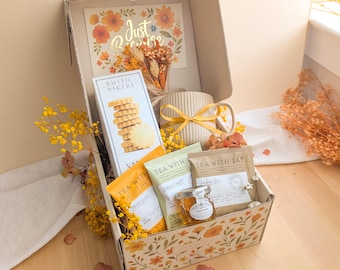 May include: A gift box containing a Rustic Bakery vanilla shortbread box, a ribbed ceramic mug with a yellow ribbon, three Tea With Tae tea packets, and a small jar of honey. The box features a floral print and a Just Because card with dried flowers.