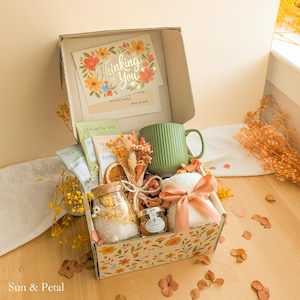 May include: A gift box containing a green ribbed ceramic mug, a glass jar of bath salts, a small jar of honey, tea packets, a white plush item tied with a peach ribbon, and a dried floral bouquet. A card inside reads Thinking of You.