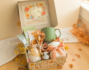May include: A gift box containing a green ribbed ceramic mug, a glass jar of bath salts, a small jar of honey, tea packets, a white plush item tied with a peach ribbon, and a dried floral bouquet. A card inside reads Thinking of You.