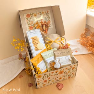 May include: A gift box containing a box of Rustic Bakery cookies, a ribbed ceramic mug with a yellow ribbon, three Tea With Tae tea pouches, and a small jar of honey. The box features a floral print and includes a thank you card and dried flowers.