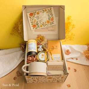 May include: A gift box containing a ceramic mug, a bar of handmade soap, a yellow tea canister, a small jar, and a yellow chocolate bar. A thank you card with floral illustrations sits inside the lid. The box is decorated with dried yellow flowers.