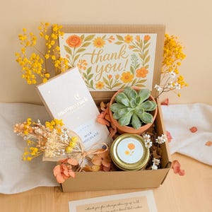 May include: A gift box containing a succulent in a terracotta pot, a chocolate bar, and a jar with a floral design. A card with the words "thank you" is also included. The box is decorated with dried flowers and greenery.