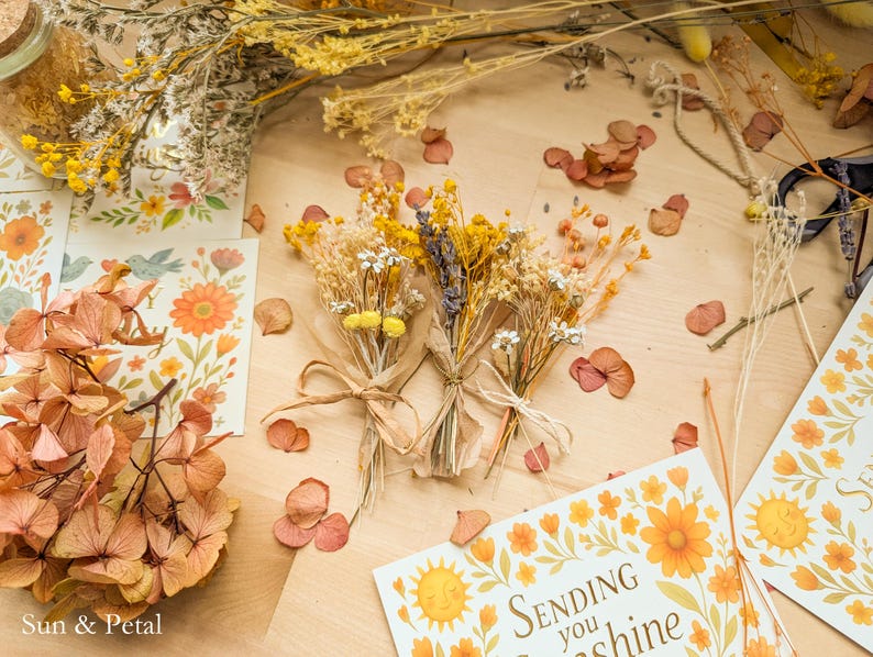 May include: Three small bouquets of dried yellow and white flowers tied with twine, surrounded by loose dried petals, floral cards with the text "Sending you sunshine," and a glass jar filled with yellow dried flowers on a light wood surface.