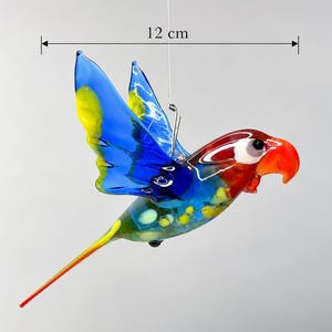 Könnte beinhalten: Ein hängendes Papageien-Ornament aus Glas in leuchtenden Blau-, Gelb-, Rot- und Grüntönen. Der Vogel hat einen gebogenen orangefarbenen Schnabel und einen langen Schwanz. Eine Messlinie zeigt eine Breite von 12 cm an. Die Glasfigur hängt an einem Draht.