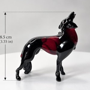 Può includere: Una statuetta in vetro raffigurante un lupo che ulula, caratterizzata da un esterno nero lucido con accenti rosso scuro traslucidi sul corpo. La scultura è alta 8,5 cm, come indicato dal testo di misurazione visibile a sinistra.