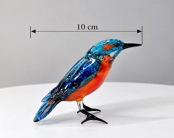 Op de afbeelding: Een glazen vogelbeeldje met een levendig blauw en oranje kleurenpalet met een gespikkeld patroon op de vleugels en rug. De vogel heeft een zwarte snavel en dunne zwarte pootjes. Een meetlijn geeft een totale lengte van 10 cm aan.