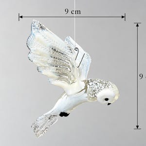 Könnte beinhalten: Ein Eulen-Ornament aus klarem und weißem Glas in Flugpose mit strukturierten Flügeln und einem gepunkteten Kopfmuster. Die Figur hat einen kleinen schwarzen Schnabel und Augen, mit sichtbaren Maßlinien, die eine Breite und Höhe von 9 cm anzeigen.
