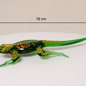 Könnte beinhalten: Eine grüne Glas-Eidechsenfigur mit orangefarbenen und gelben Flecken auf dem Rücken und Kopf. Die Eidechse ist horizontal positioniert, und eine Messanzeige darüber zeigt eine Gesamtlänge von 16 cm. Das Glas hat eine glatte, glänzende Oberfläche.