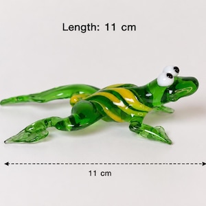 Op de afbeelding: Een groen glazen kikkerbeeldje met gele strepen op de rug en witte ogen met zwarte pupillen. De kikker staat in een springende houding en is 11 cm lang, zoals aangegeven door de tekst en de meetpijl in de afbeelding.