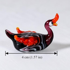 May include: A small glass figurine of a bird with a long neck, featuring a black, white, and orange marbled pattern. The bird has a bright orange crest and beak, with white and black eyes. It measures 1.57 inches in length as indicated by text.