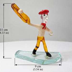 Könnte beinhalten: Eine Glasfigur einer Person mit rotem Barett und rotem Halstuch, gelber Hose und schwarzen Schuhen, die einen gelben Schläger und einen weißen Ball hält. Die Figur steht auf einem klaren Glassockel und ist 11 cm hoch und 9 cm breit.