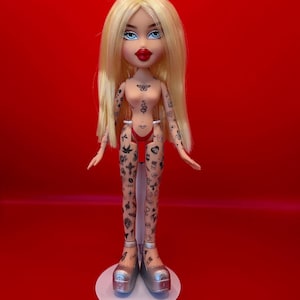 May include: A Bratz doll with long blonde hair, blue eyes, and red lipstick. The doll has multiple tattoos on its arms, torso, and legs. It is wearing a red bikini and silver platform shoes. The doll is standing on a white base against a red background.