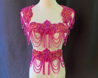 May include: A two-piece costume set made of pink beads and sequins, featuring a halter top and a belt. The top has a floral design and the belt has a similar design.